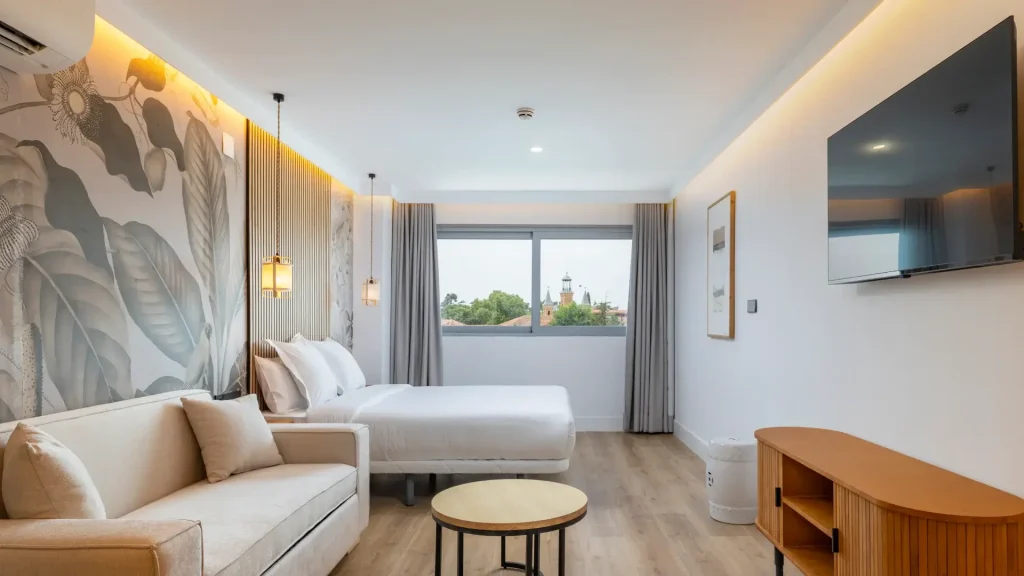 Habitación moderna con cama, sofá, mesa y televisión, decorada con murales y luz suave.