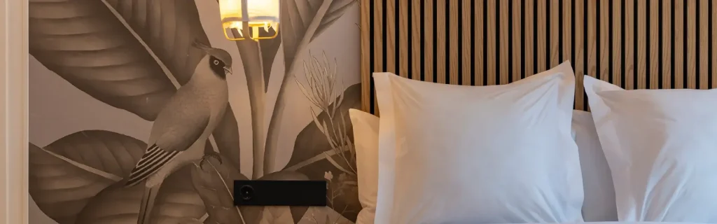 Cama con cabecero de madera, paredes con mural de ave y hojas, y luminaria moderna.