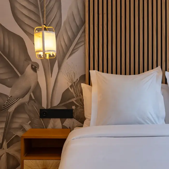 Cama con cabecera de madera, lámpara colgante y mural de hojas y pájaro en blanco y negro.