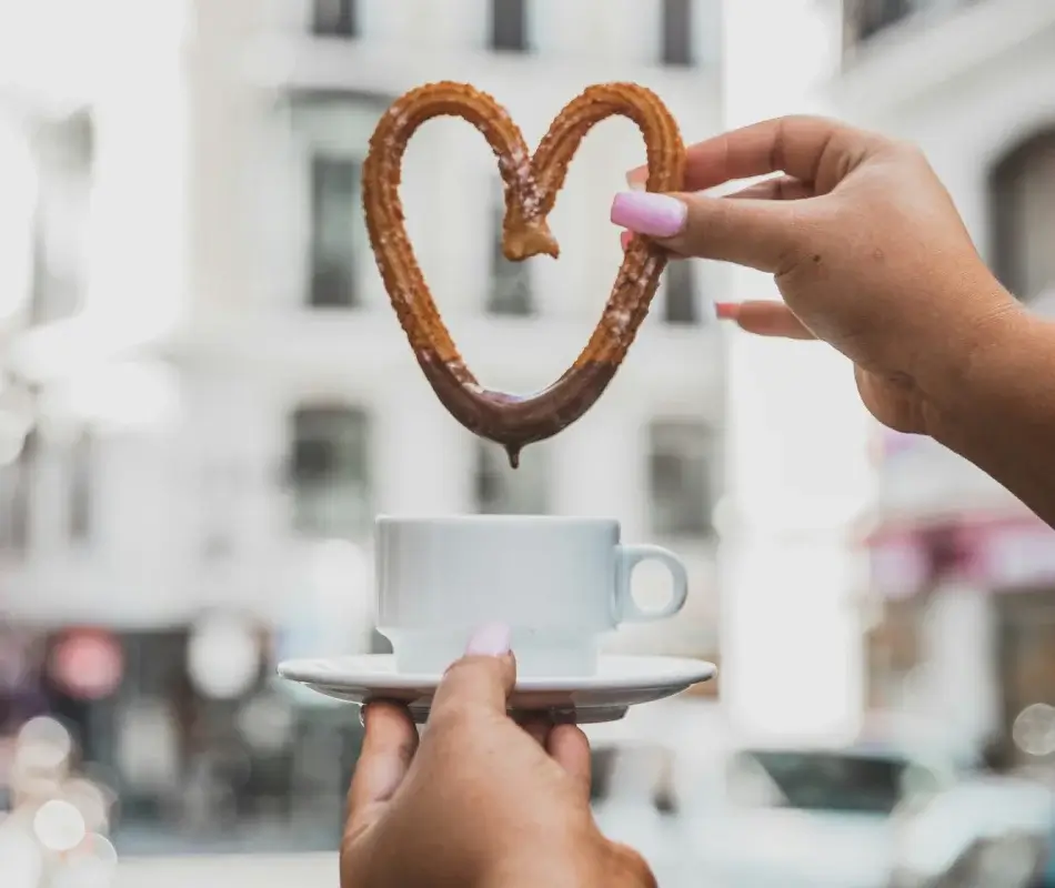 Manos sostienen churro en forma de corazón sobre taza blanca, con fondo urbano desenfocado.