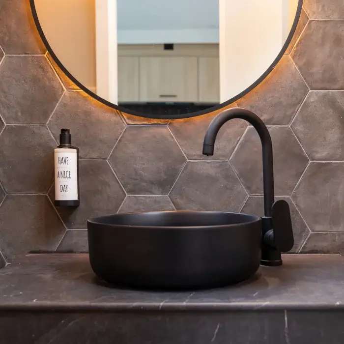 Lavabo negro sobre encimera gris con pared de azulejos hexagonales y espejo redondo iluminado.