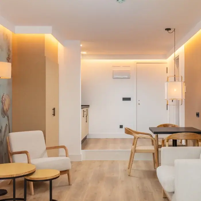 Sala moderna con muebles de madera, sofá blanco y cocina integrada al fondo, iluminación cálida.