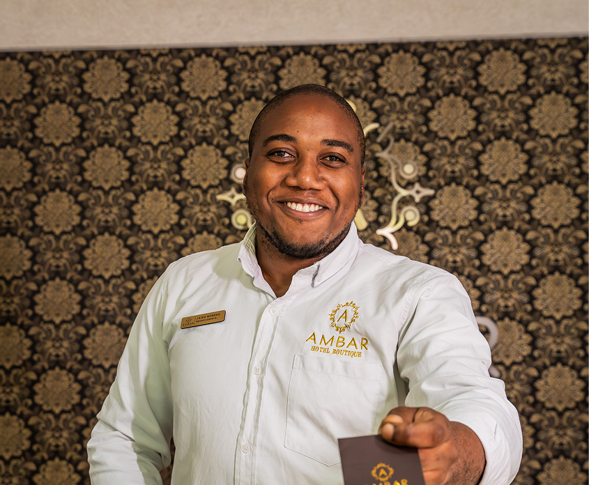 Persona sonriente con camisa blanca de "Ámbar Hotel Boutique" extendiendo una tarjeta, fondo decorativo elegante.