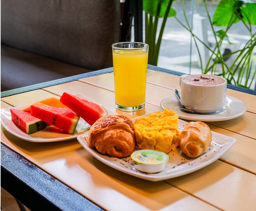 Desayuno con fruta, jugo, café, huevos revueltos, croissant y pan en una mesa junto a una ventana.