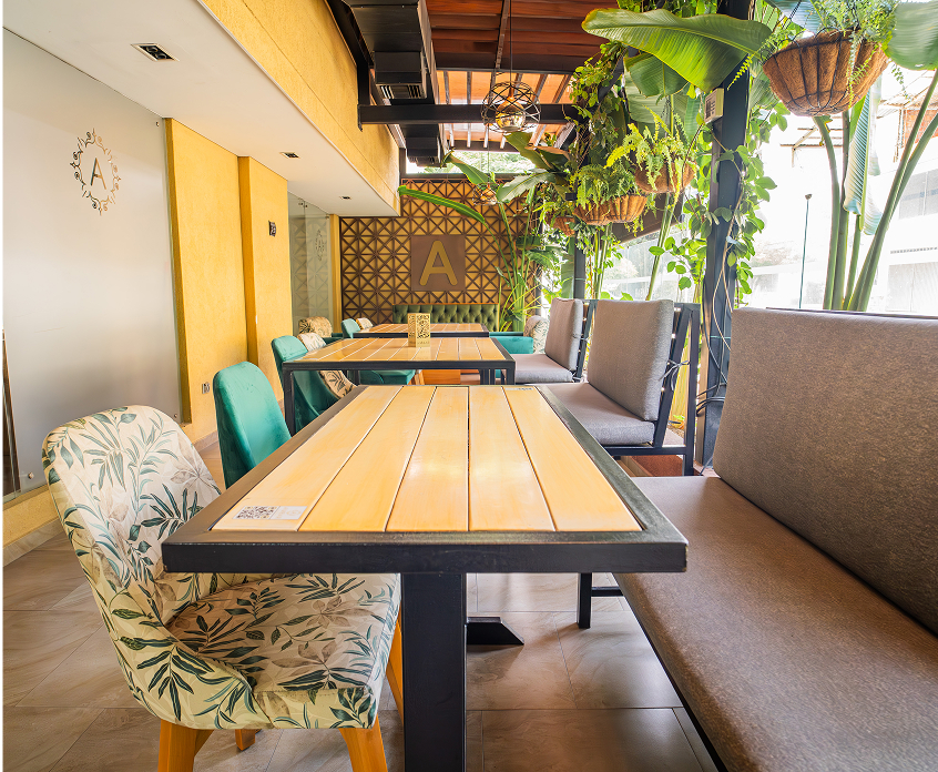 Interior de un restaurante con mesas de madera, sillas decoradas y plantas colgantes, creando un ambiente acogedor.