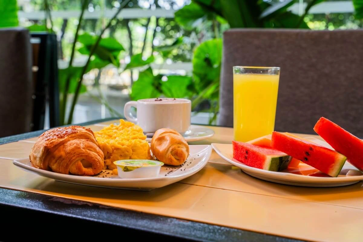 Desayuno con croissants, huevos revueltos, jugo de naranja, rodajas de sandía y taza de café en mesa exterior.