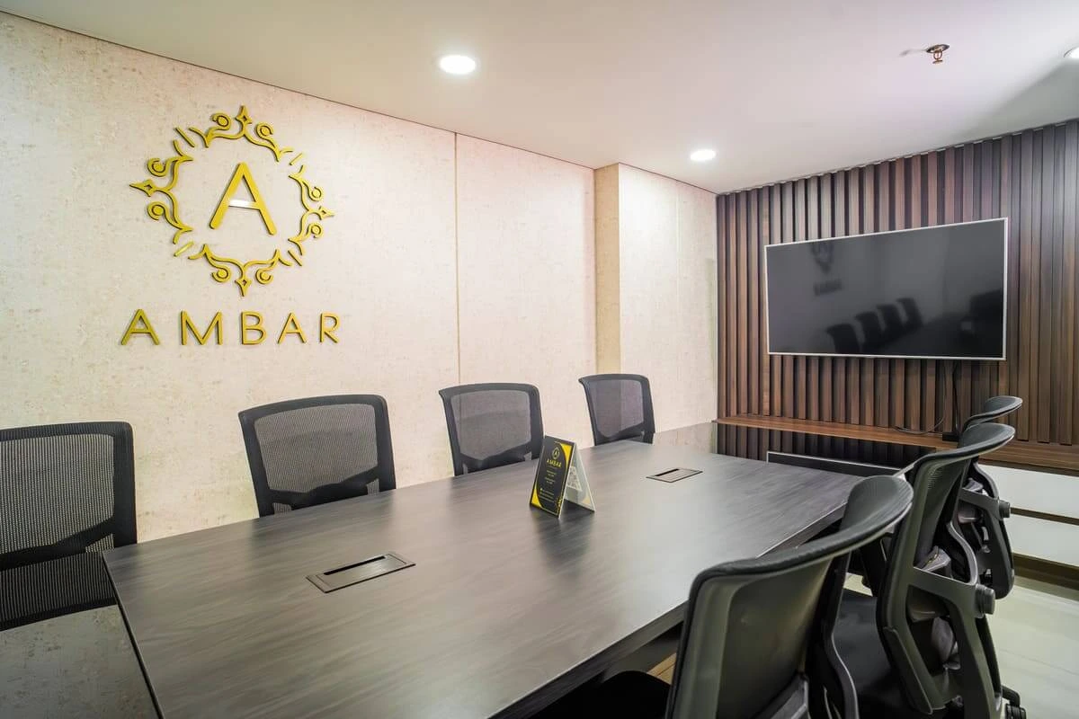 Sala de conferencias moderna con mesa larga, sillas negras, pantalla grande y el logotipo "AMBAR" en la pared.