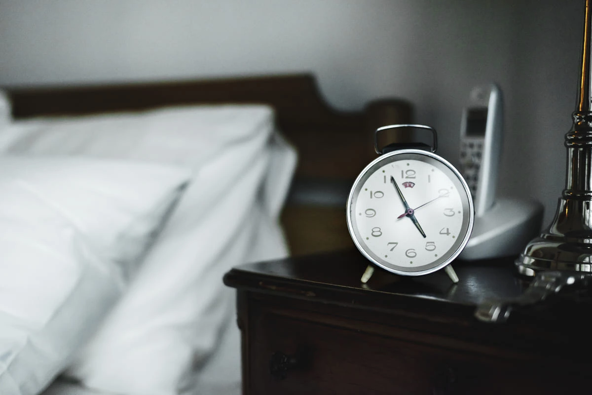 Despertador plateado en una mesita junto a una cama. Al fondo, un teléfono inalámbrico y una lámpara metálica.