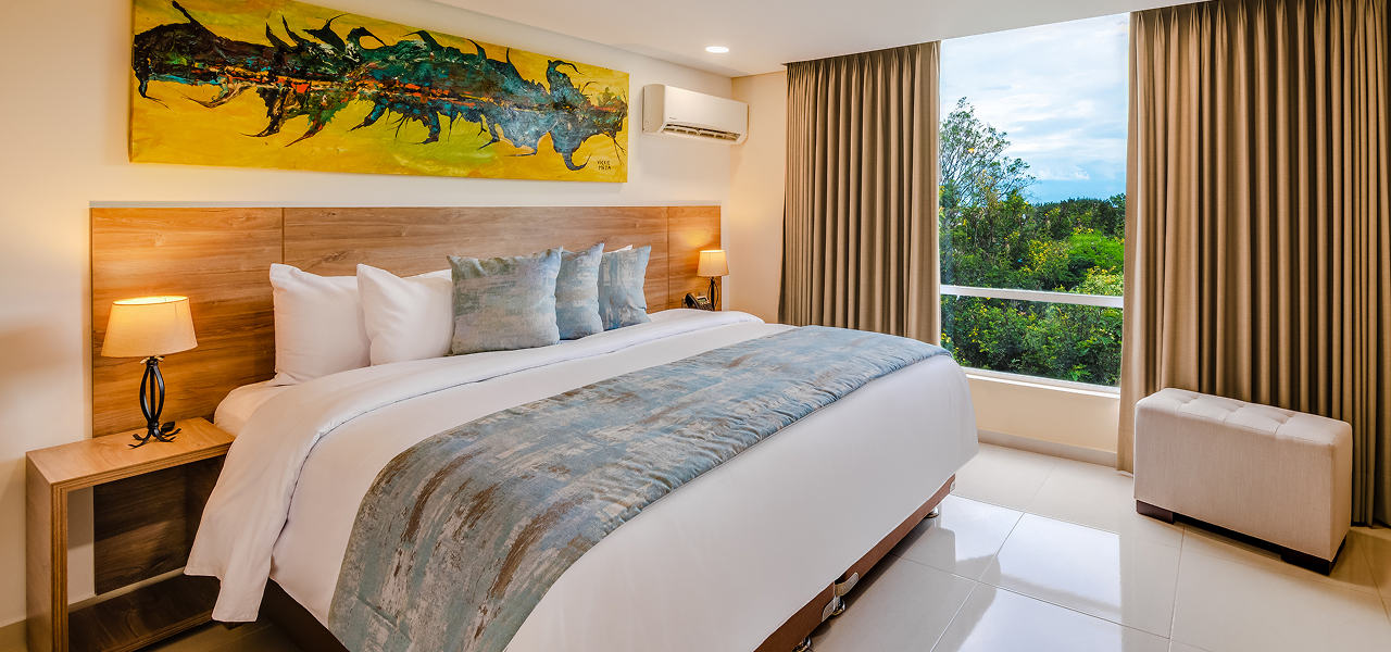 Habitación luminosa con cama grande, pintura en la pared, ventanas con vista al exterior y cortinas beige.