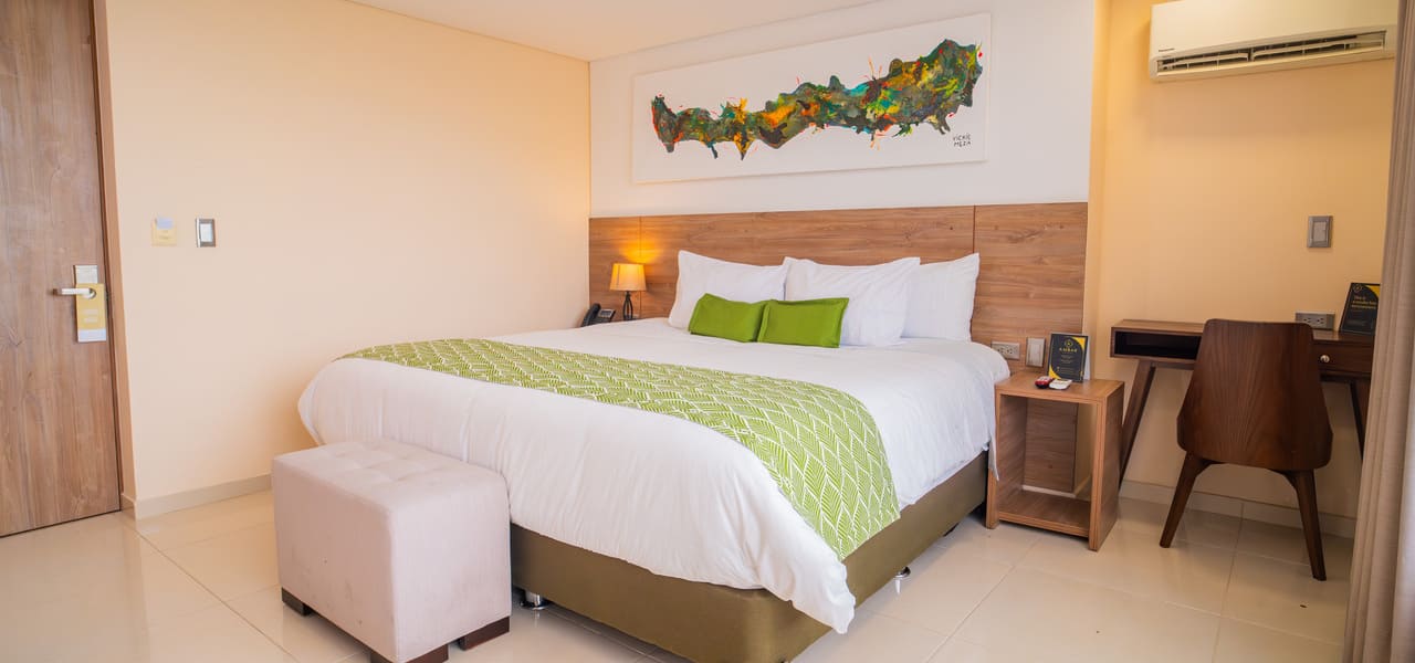 Habitación de hotel moderna con cama grande, cojines verdes, escritorio, arte en la pared y aire acondicionado.