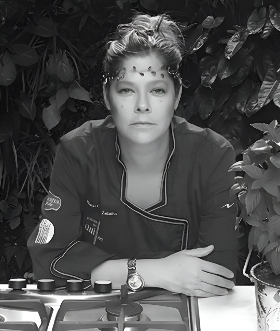 Mujer con uniforme de chef y peinado creativo, frente a una estufa, rodeada de plantas en un ambiente oscuro.