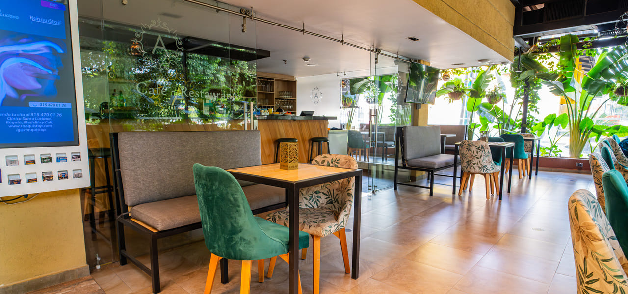Cafetería moderna con mesas y sillas coloridas, rodeada de plantas, con ventanales que dejan entrar luz natural.