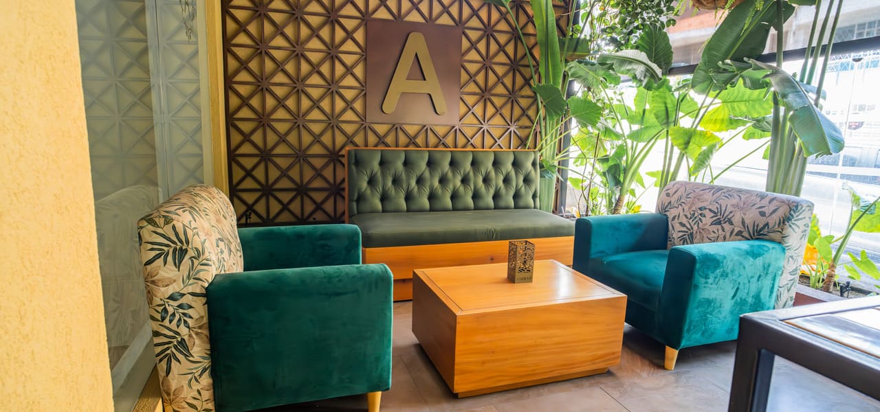 Sala elegante con sillones verdes, mesas de madera y una pared decorativa con la letra "A" y plantas alrededor.