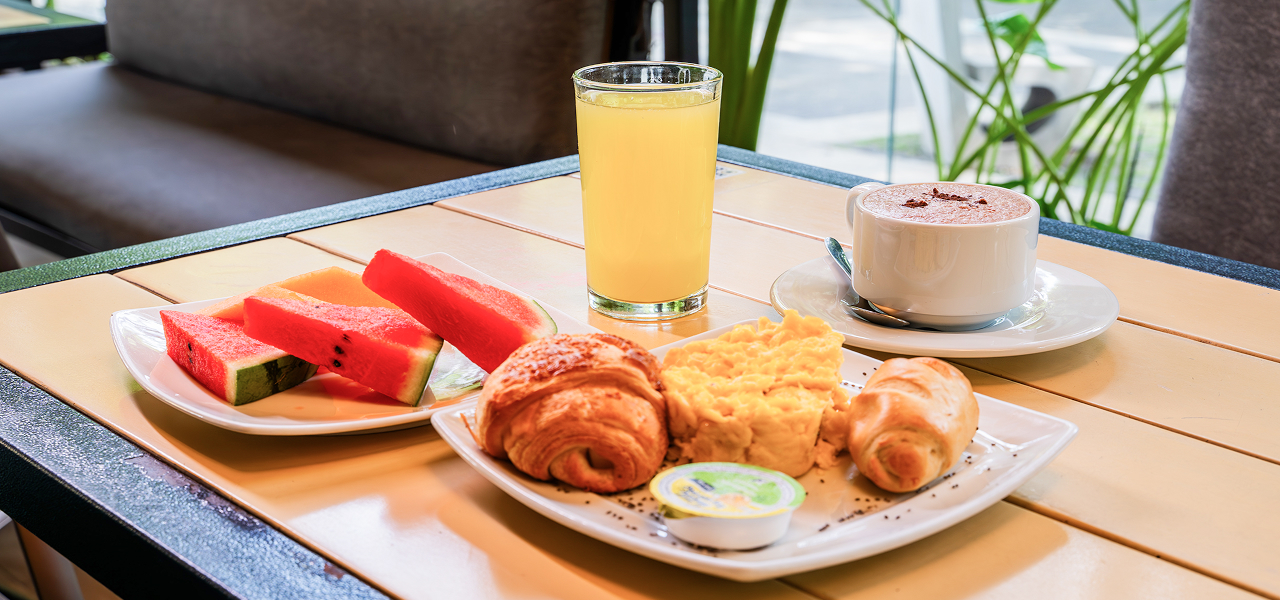 Desayuno con café, jugo, croissants, huevos revueltos y fruta en un plato sobre una mesa de madera.