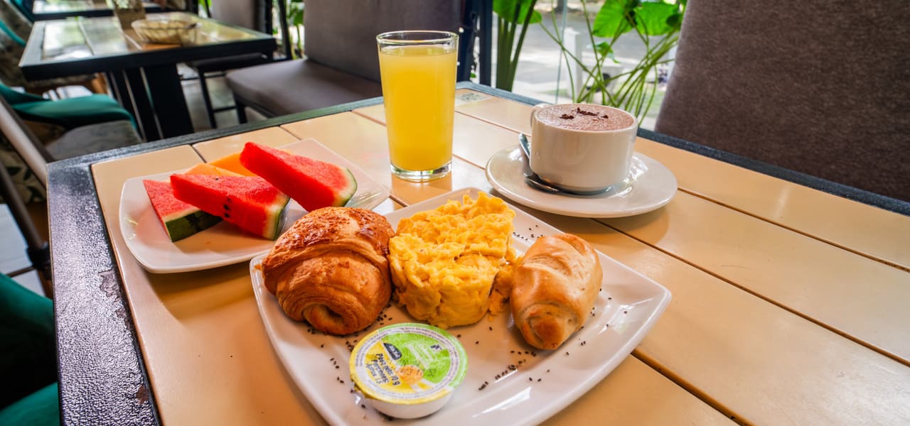 Desayuno en mesa: huevos revueltos, croissants, fruta, jugo de naranja y café al aire libre.