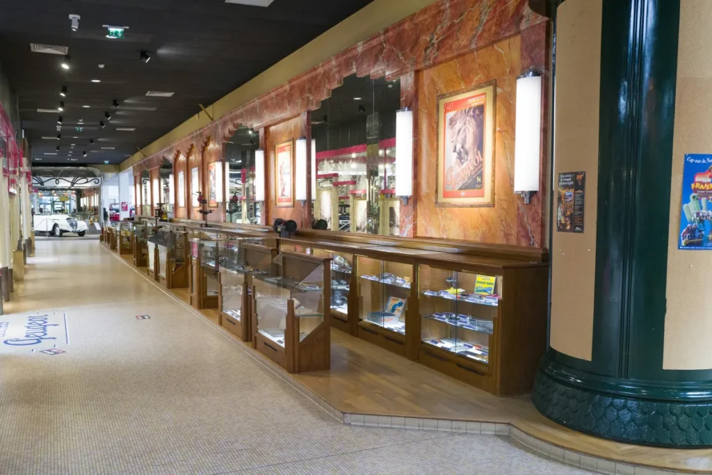 Un couloir de musée avec des vitrines en bois et verre, posters muraux et une voiture blanche en fond.