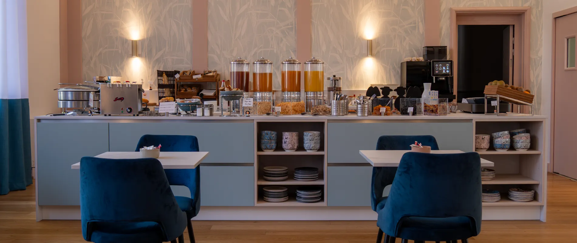 Buffet de petit-déjeuner avec jus, céréales et pain, entouré de chaises bleues, décor moderne et chaleureux.