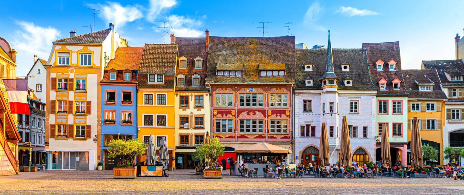 Façades colorées de maisons traditionnelles alignées sur une place animée avec cafés en plein air, ciel bleu.