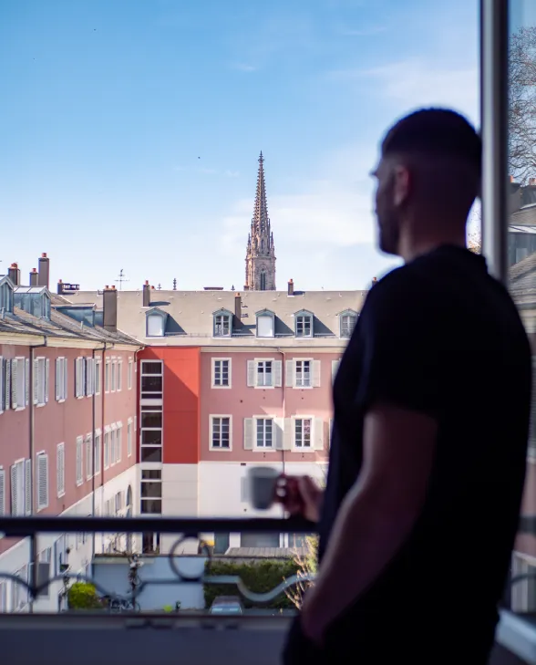 Homme regardant une cathédrale depuis une fenêtre, tenant une tasse, journée ensoleillée.