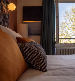 Chambre avec lit douillet, coussins orange et fenêtre ensoleillée avec vue sur des arbres.