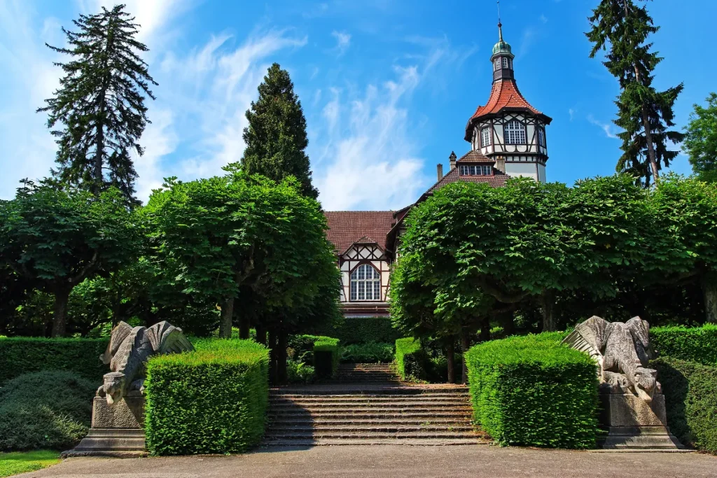 Bâtiment à colombages avec tour, encadré par des arbres, escalier et statues de créatures ailées au premier plan.