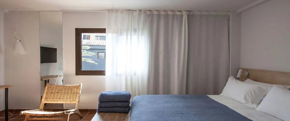 Habitaci&oacute;n minimalista con cama, silla de mimbre y ventana con cortinas blancas.