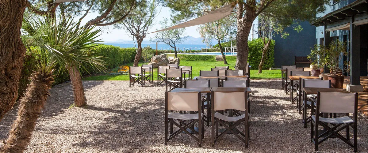 Terraza con mesas y sillas de madera, rodeada de &aacute;rboles y con fondo de c&eacute;sped iluminado.