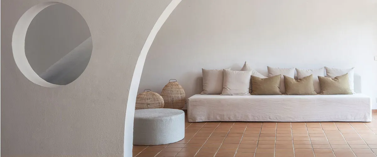 Sala minimalista con sof&aacute; blanco, cojines y cestas frente a pared blanca con arco y ventana circular.