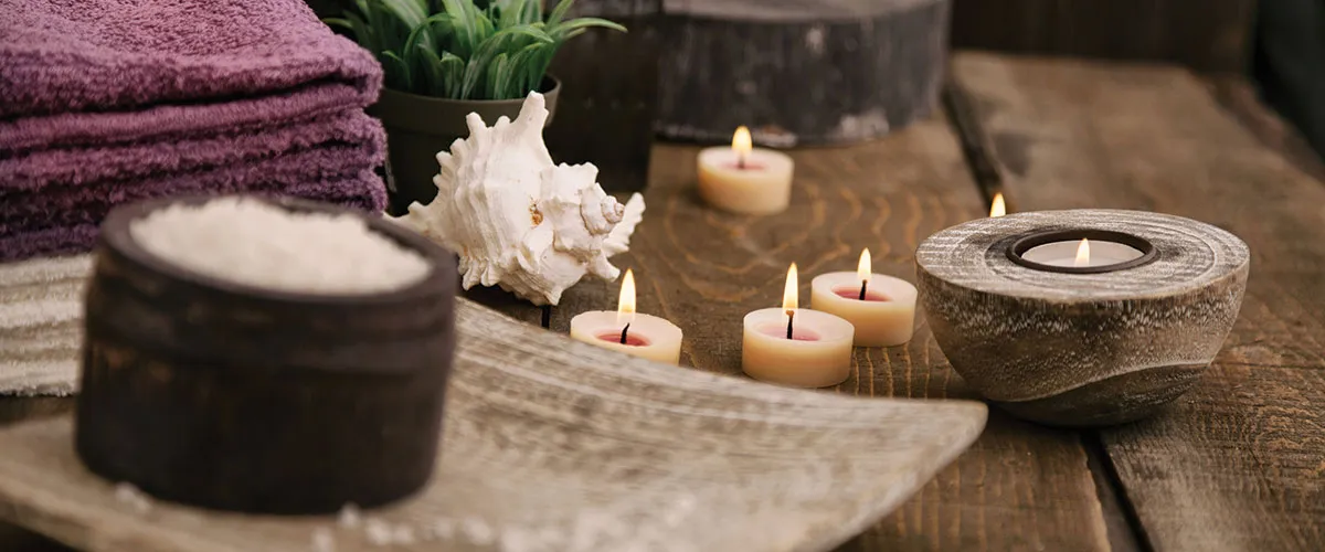 Velas encendidas sobre mesa de madera, toallas moradas, sales de ba&ntilde;o y caracola decorativa en ambiente relajante de spa.