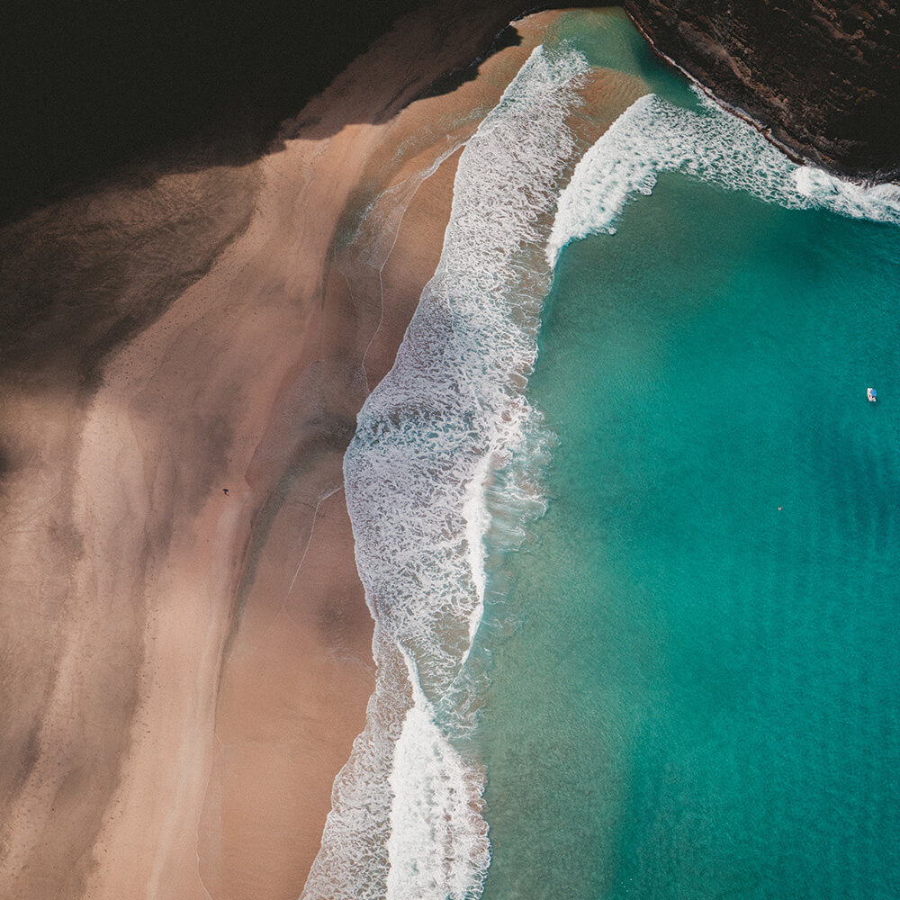 Vista a&eacute;rea de una playa con arena clara, olas blancas y mar turquesa brillante, bordeada por formaciones rocosas oscuras.