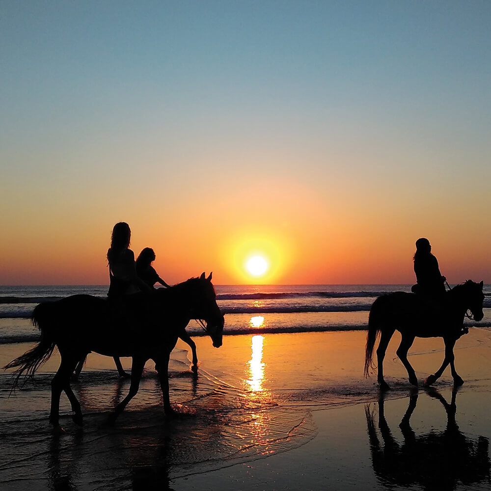 Personas a caballo por la playa al atardecer, con el sol reflej&aacute;ndose en el agua.