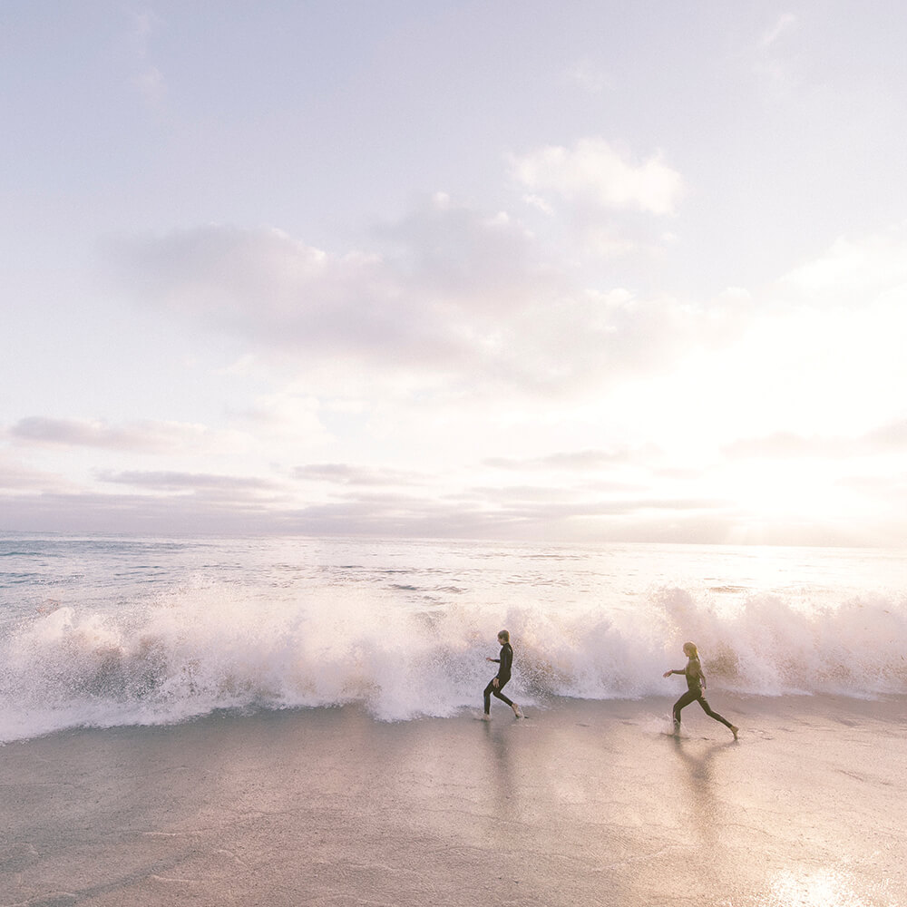 Dos personas corren junto al mar, bajo un cielo nublado y con olas rompiendo en la orilla al atardecer.