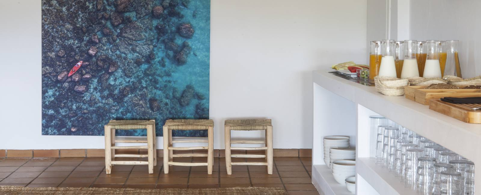 Interior minimalista con bancos de madera, mural de un kayak y mesa con jugos y vasos en estantes.