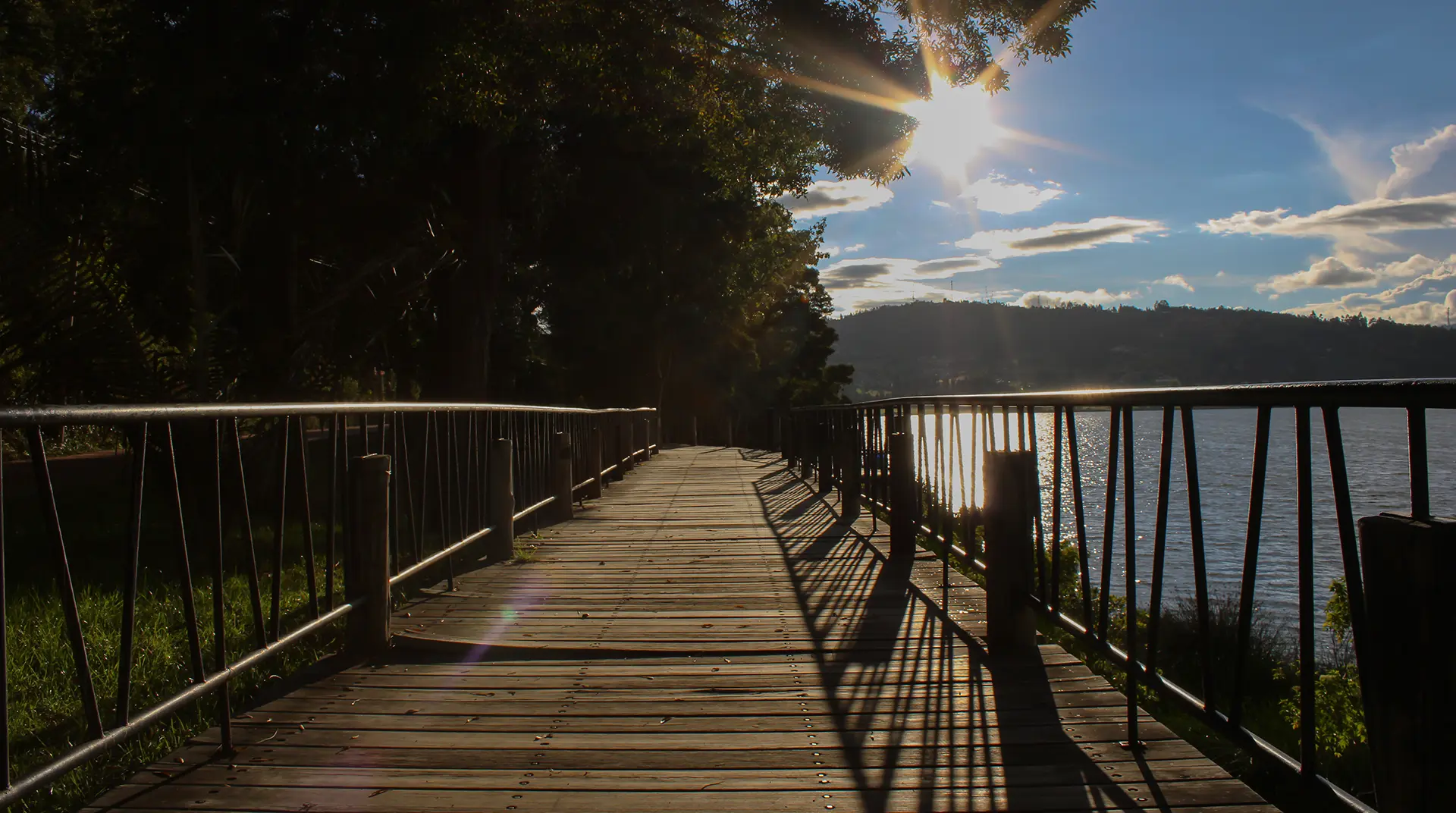 Pasarela de madera al atardecer junto a un lago, con árboles a la izquierda y sol reflejándose en el agua.
