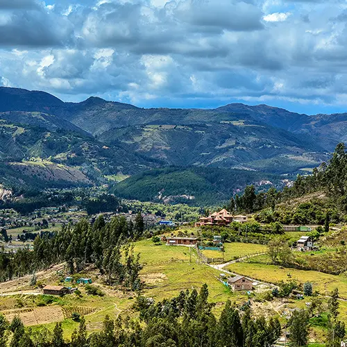 Paisaje montañoso con praderas, árboles y casas dispersas bajo un cielo parcialmente nublado.
