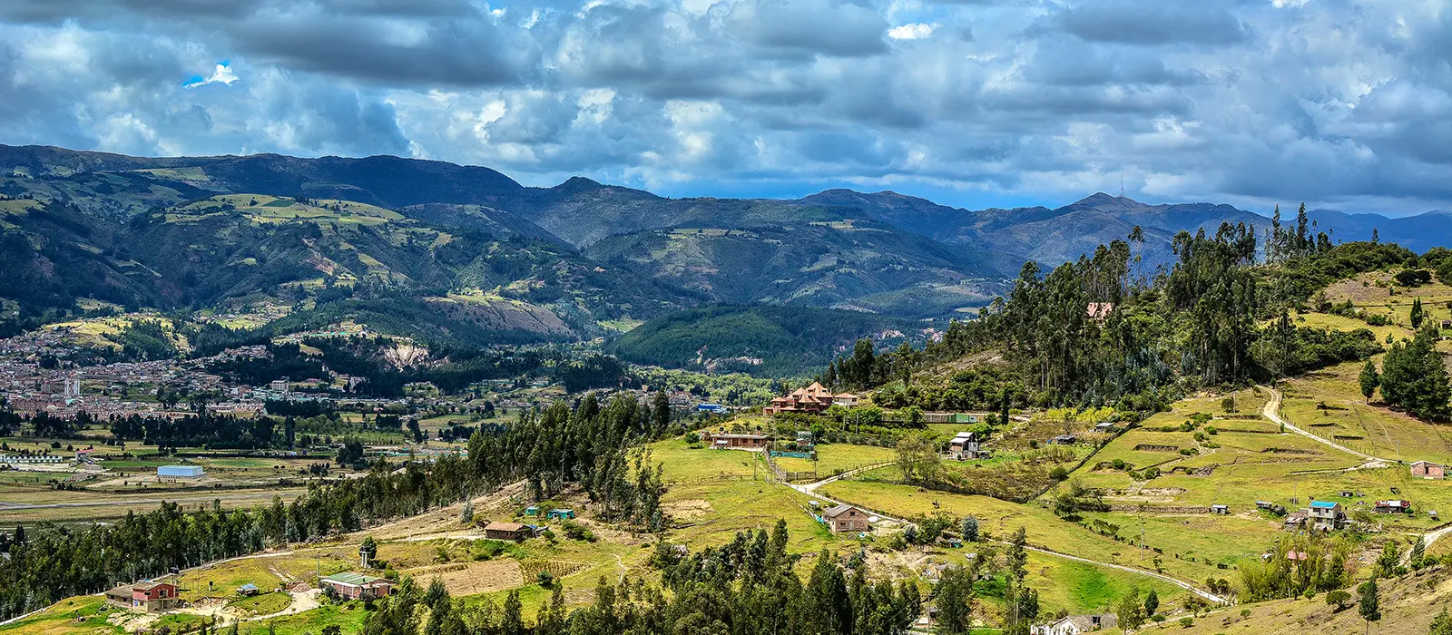 Valle verde con montañas al fondo, pequeñas casas y caminos serpentean entre campos y árboles bajos bajo un cielo nublado.