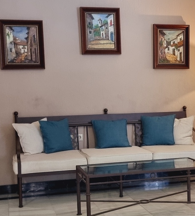 Sofá con cojines azules y crema frente a mesa de vidrio, con tres cuadros de paisajes urbanos en la pared beige.