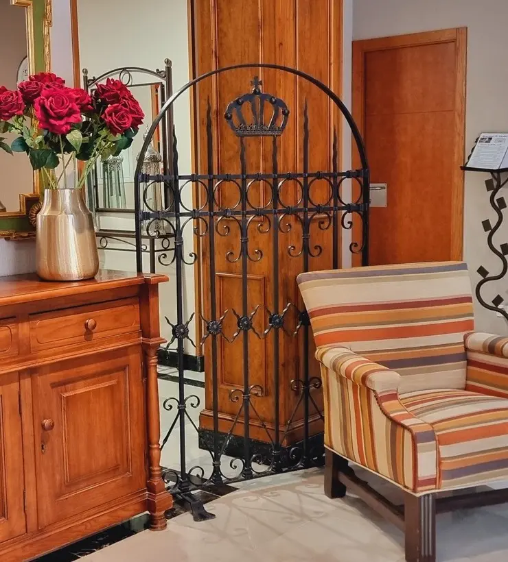 Mueble de madera con jarrón dorado y rosas, reja con corona, y silla a rayas en habitación elegante.