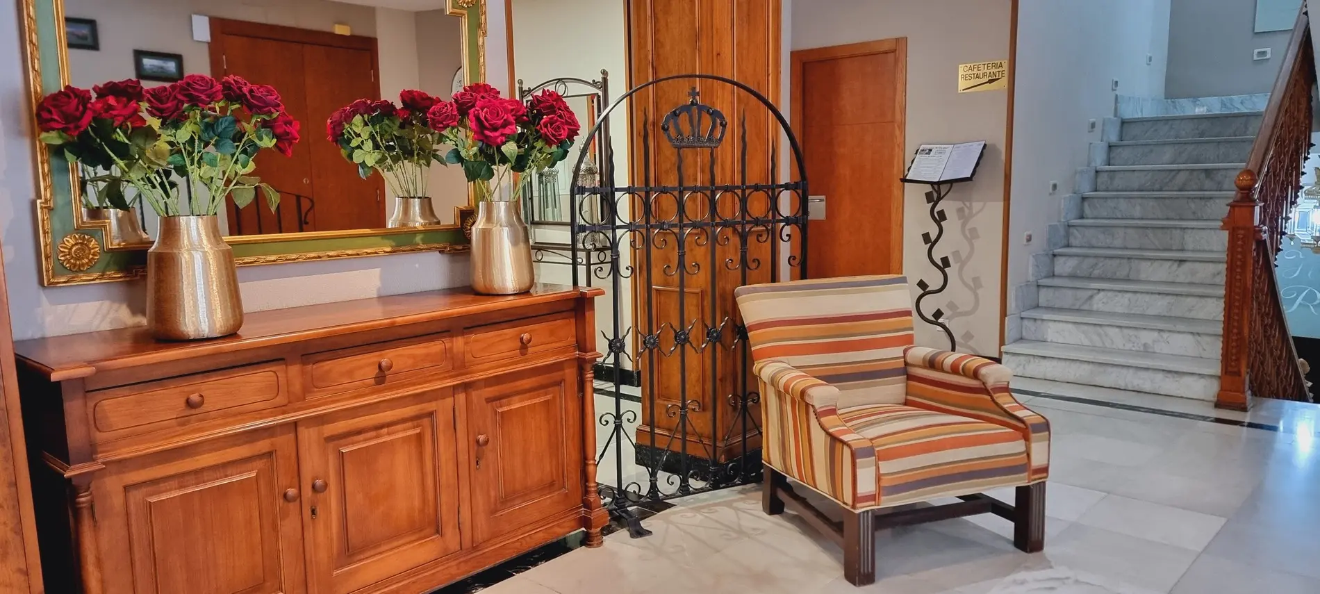 Vestíbulo con espejo, jarrones con rosas rojas, sillón a rayas y escalera de mármol. Señal de cafetería.