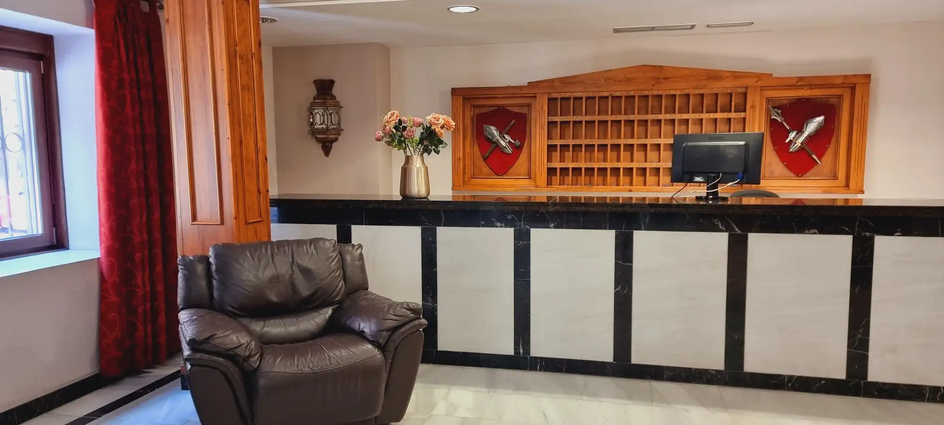 Recepción de hotel con mostrador oscuro, sillón marrón, flores en jarrón dorado, escudos en la pared y computadora.