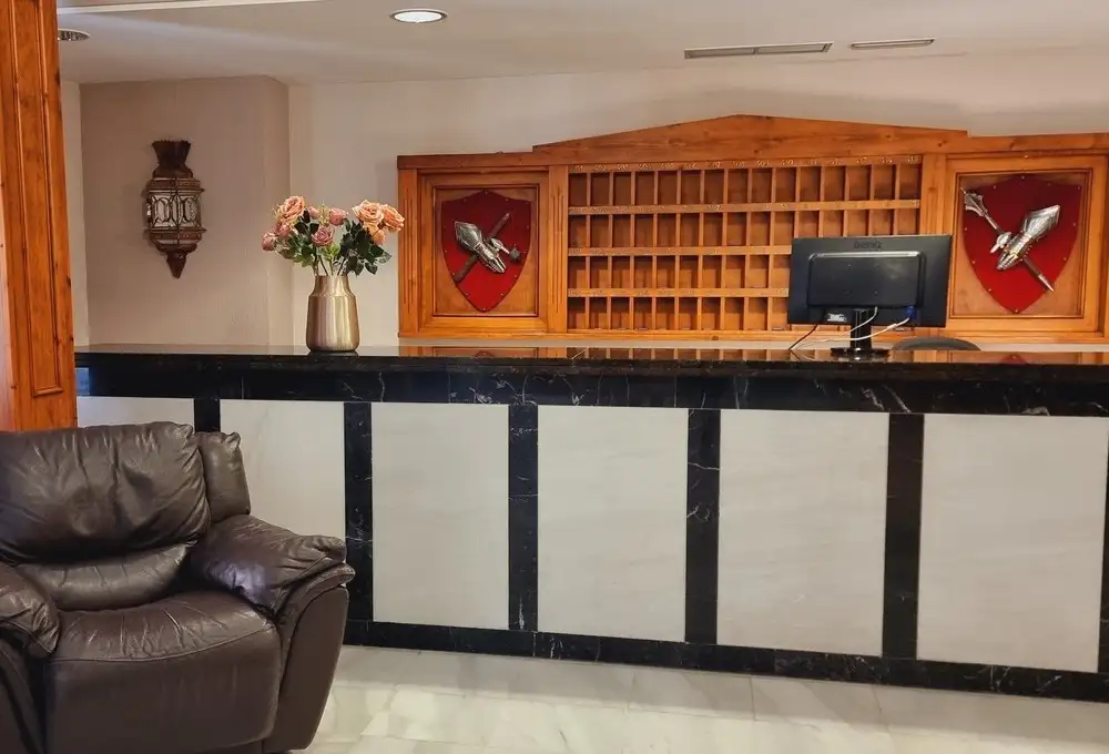 Recepción de hotel con mostrador oscuro, computadora, flores en jarrón y decoración de madera; sillón de cuero marrón.
