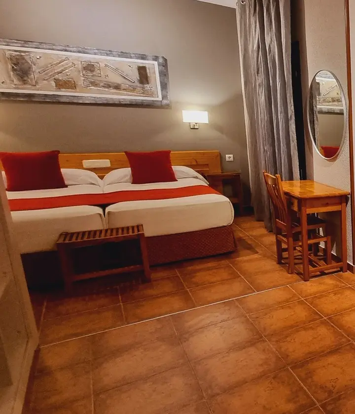 Habitación de hotel con dos camas individuales, mesa y silla de madera, lámpara de pared, cuadro y espejo redondo.