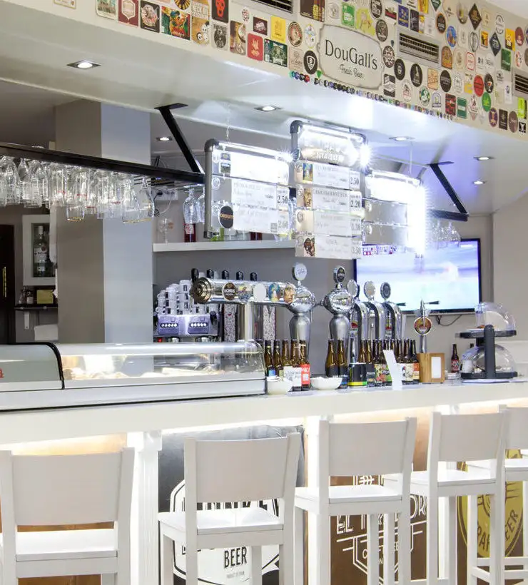 Barra de bar con taburetes blancos, grifos de cerveza, botellas y vasos. Decoración con carteles y portavasos.