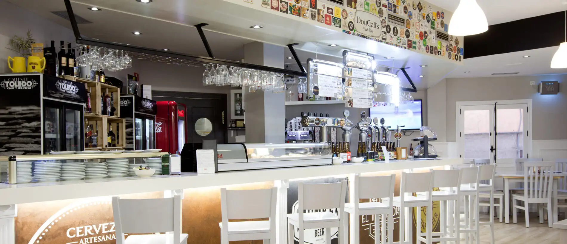 Bar moderno con barra blanca, botellas en estantes de madera, copas colgantes y carteles de cerveza en la pared.