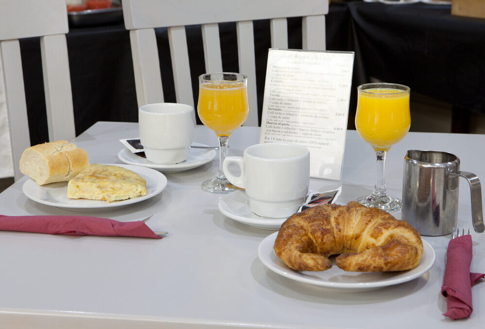 Desayuno en mesa: tortilla, croissant, café, jugo de naranja, servilletas rojas, menú al fondo.