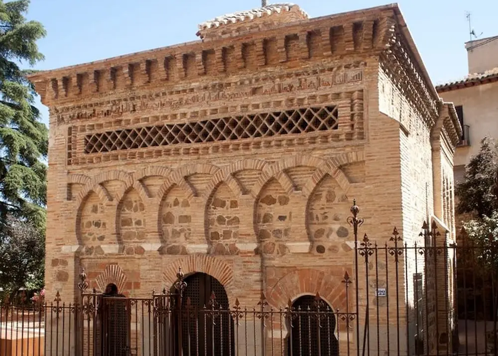 Edificio histórico de ladrillo con arcos y detalles arquitectónicos árabes, rodeado de una reja de metal.