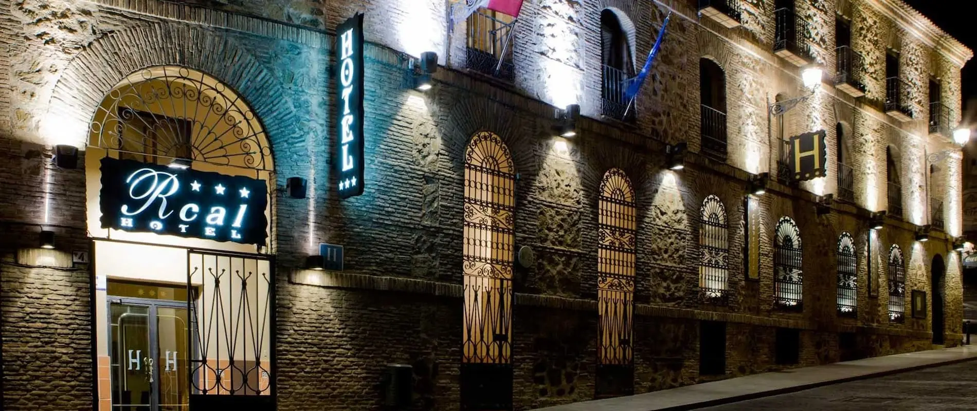 Fachada de un hotel iluminado por la noche, con arcos y letreros brillantes. Hay banderas en el edificio de ladrillo.