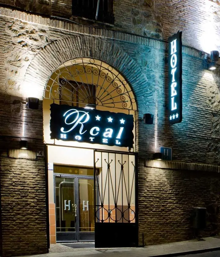 Entrada iluminada de un hotel con un cartel que dice "Rcal" y "HOTEL" con estrellas en la fachada de ladrillo.