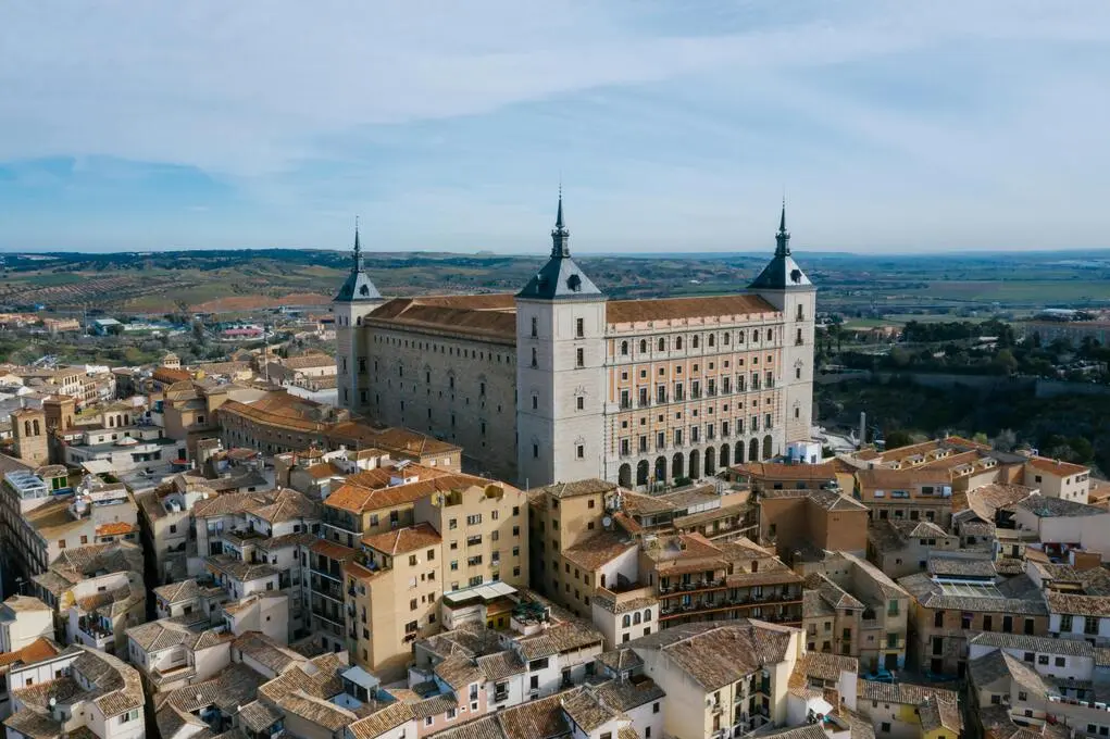 Vista aérea de un gran edificio histórico con cuatro torres, rodeado de casas con techos de tejas y un paisaje rural.