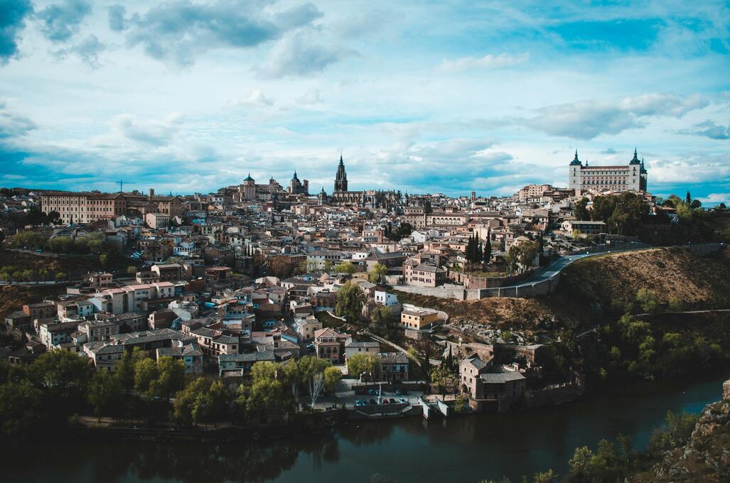 Ciudad histórica con arquitectura medieval, río en primer plano, cielo nublado.